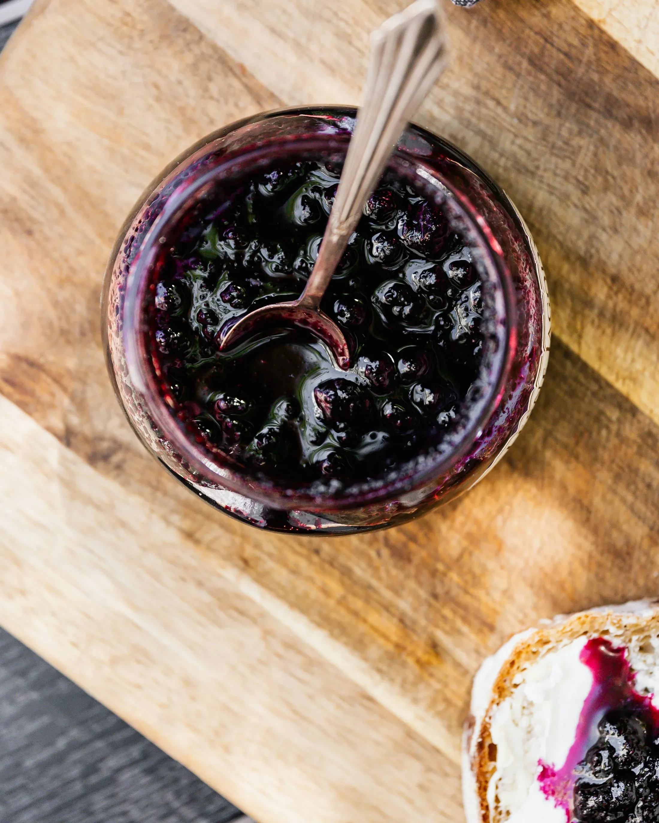 A jar of blueberry jam with a spoon resting in it on a wooden board, beside a slice of bread spread with cream cheese and jam
