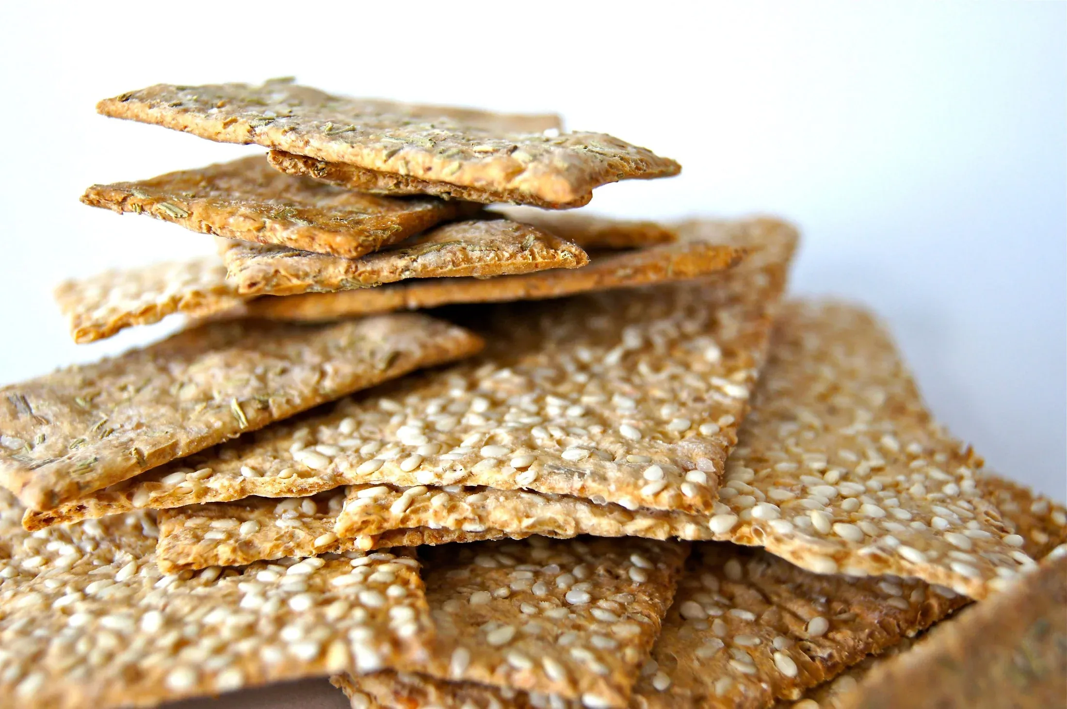 A stack of thin, pale golden seed crackers with browned edges, flecked with sesame seeds and herbs