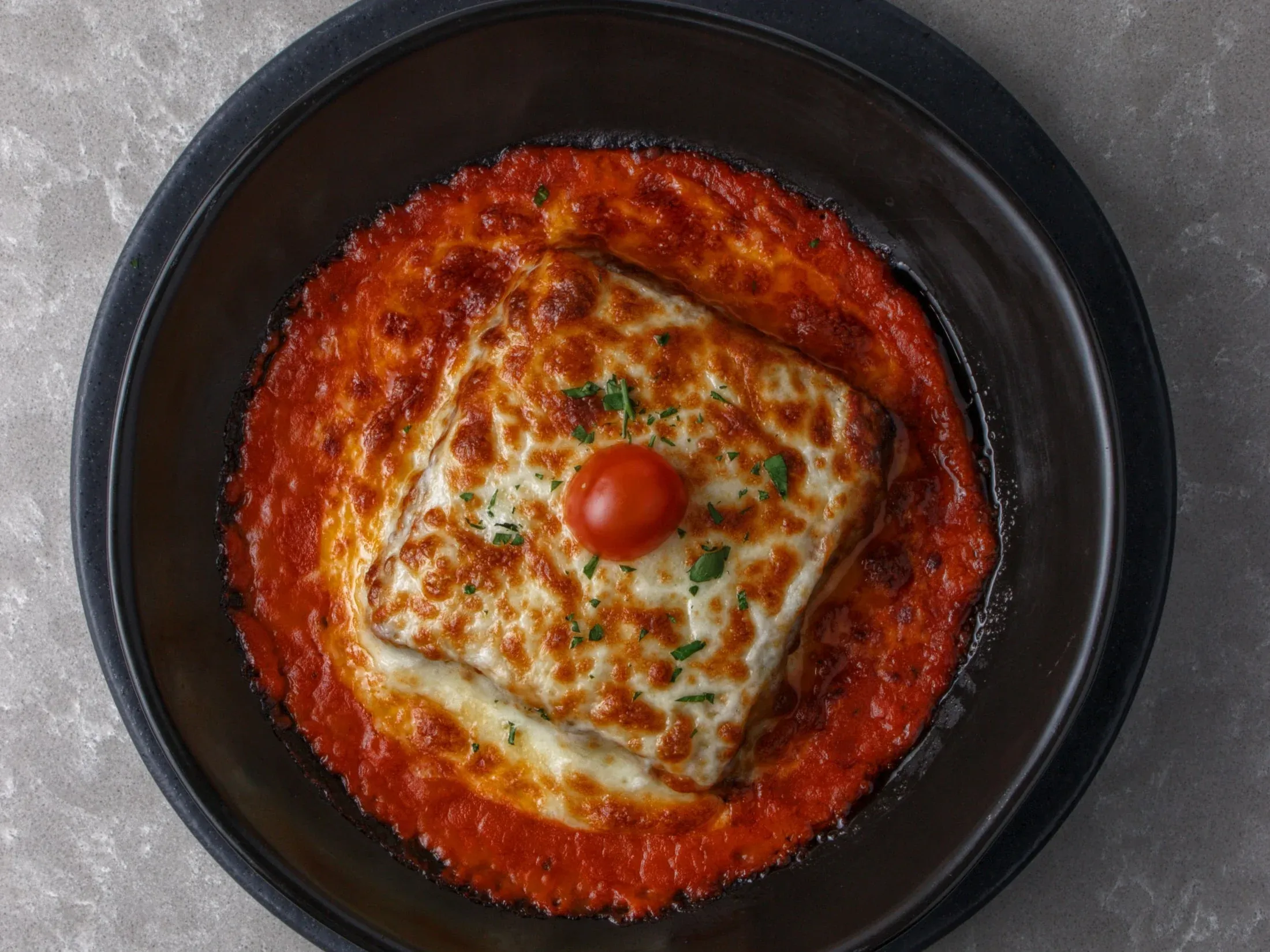 A square portion of baked lasagne topped with browned melted cheese and a cherry tomato, sitting in tomato sauce in a dark bowl